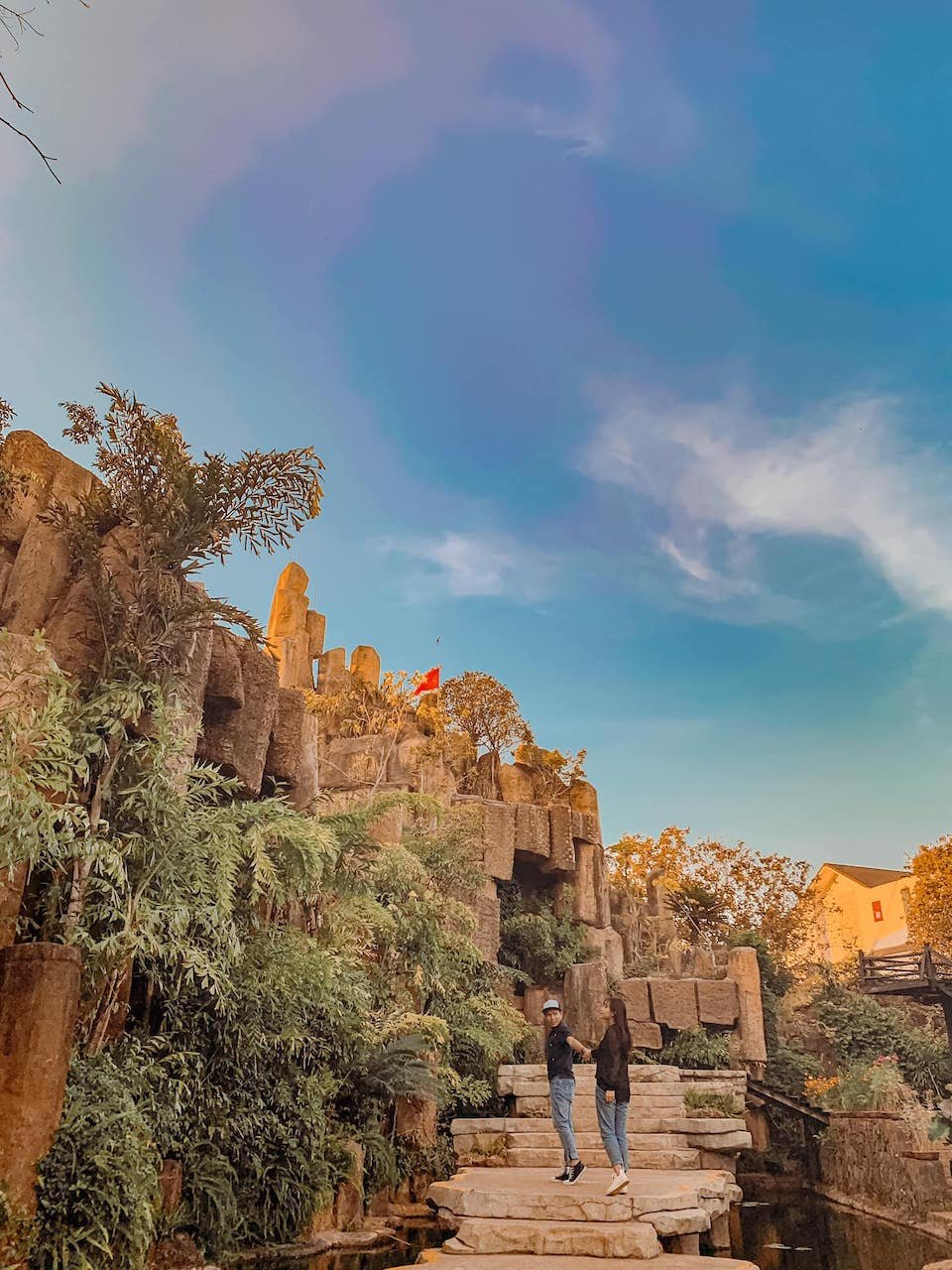 Hai người đang đứng tr&ecirc;n bậc thang đ&aacute;, xung quanh l&agrave; c&acirc;y xanh v&agrave; c&aacute;c khối đ&aacute; lớn, bầu trời xanh ph&iacute;a tr&ecirc;n.