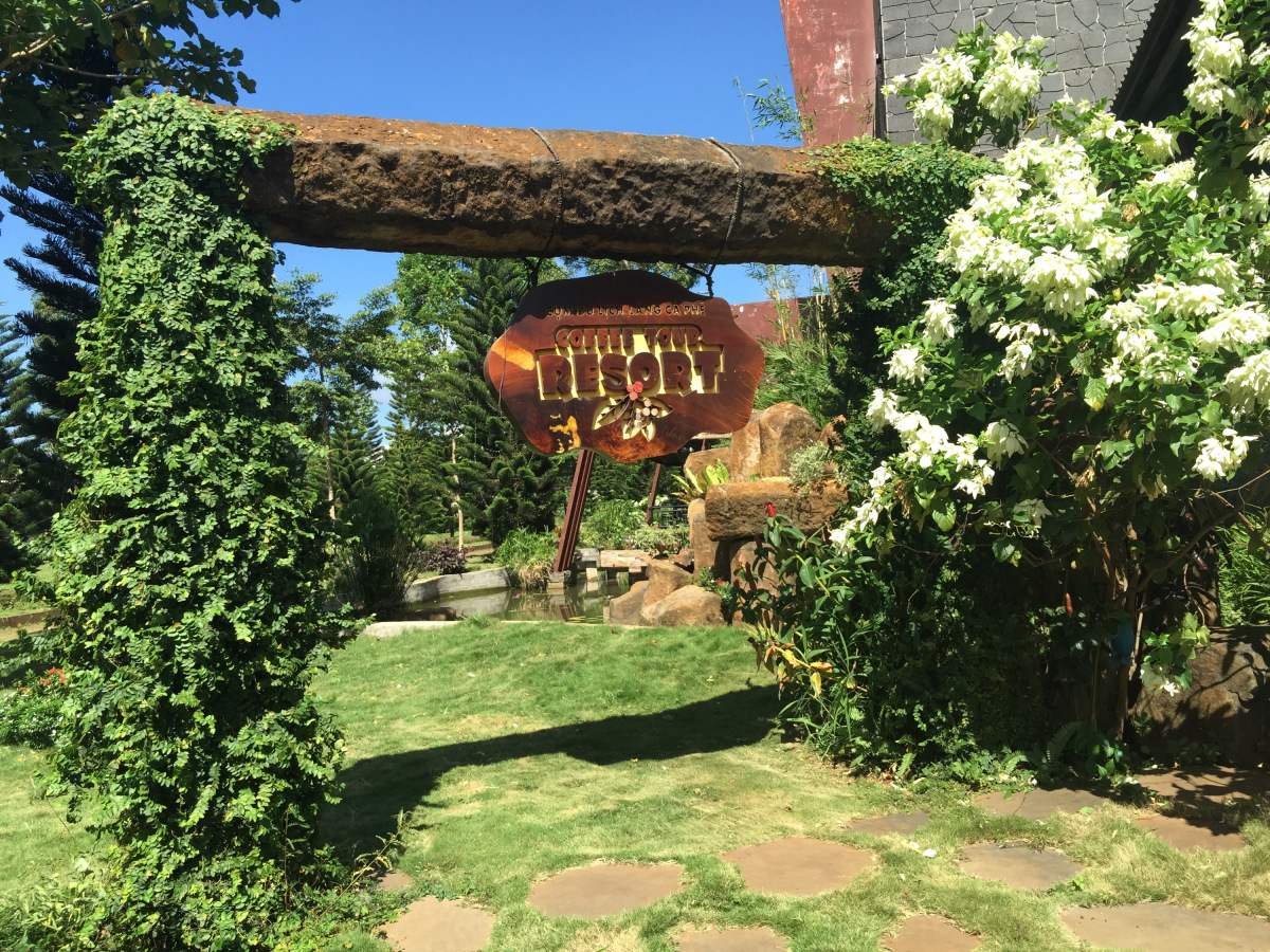 Cổng v&agrave;o khu nghỉ dưỡng với biển hiệu gỗ, xung quanh l&agrave; c&acirc;y xanh v&agrave; hoa trắng, bầu trời trong xanh.