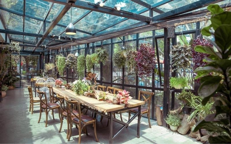 Phòng ăn trong suốt với bàn gỗ dài, ghế gỗ, cây xanh treo và ánh sáng tự nhiên từ trần kính.