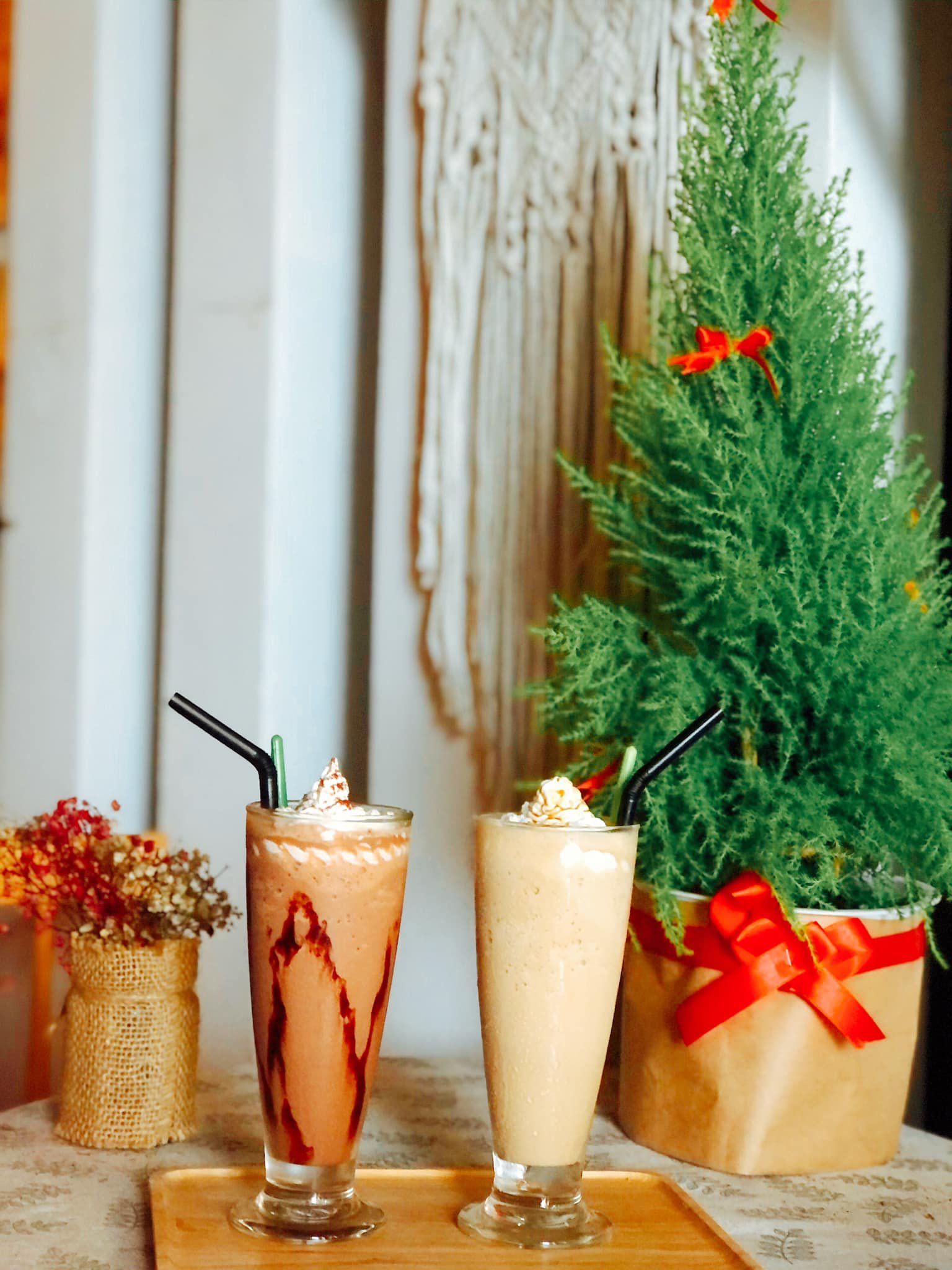 Hai ly đồ uống c&oacute; ống h&uacute;t tr&ecirc;n b&agrave;n, cạnh c&acirc;y xanh v&agrave; b&igrave;nh hoa nhỏ.