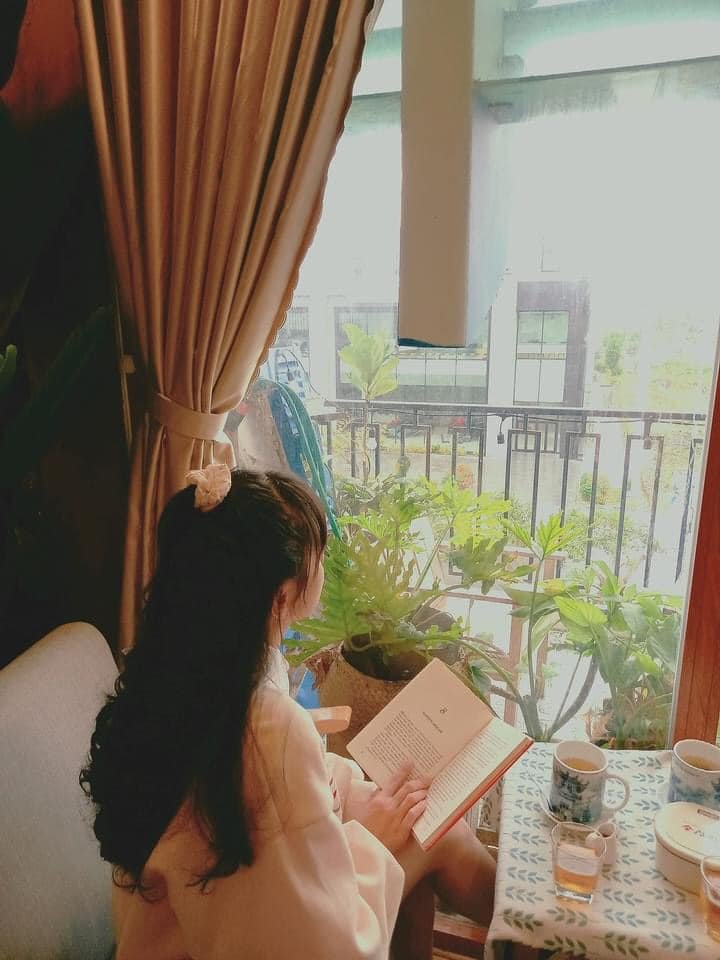 Người ngồi cạnh cửa sổ đọc s&aacute;ch, xung quanh c&oacute; c&acirc;y xanh v&agrave; b&agrave;n với cốc nước, &aacute;nh s&aacute;ng tự nhi&ecirc;n chiếu v&agrave;o.