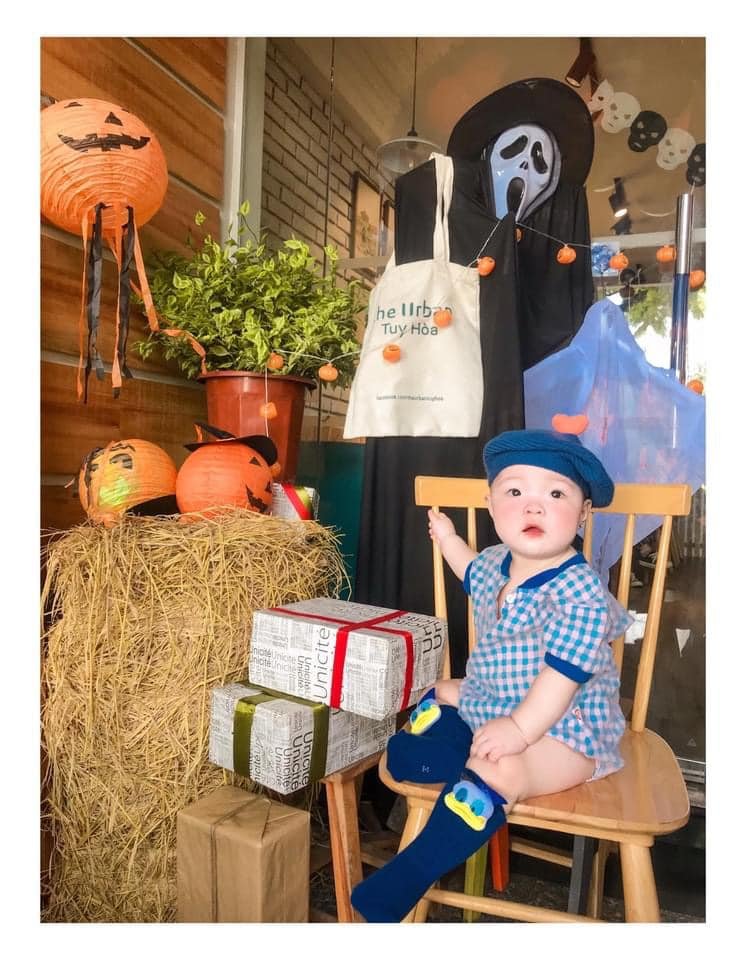 Em b&eacute; ngồi tr&ecirc;n ghế cạnh trang tr&iacute; Halloween với lồng đ&egrave;n b&iacute; ng&ocirc;, b&ugrave; nh&igrave;n v&agrave; hộp qu&agrave;.