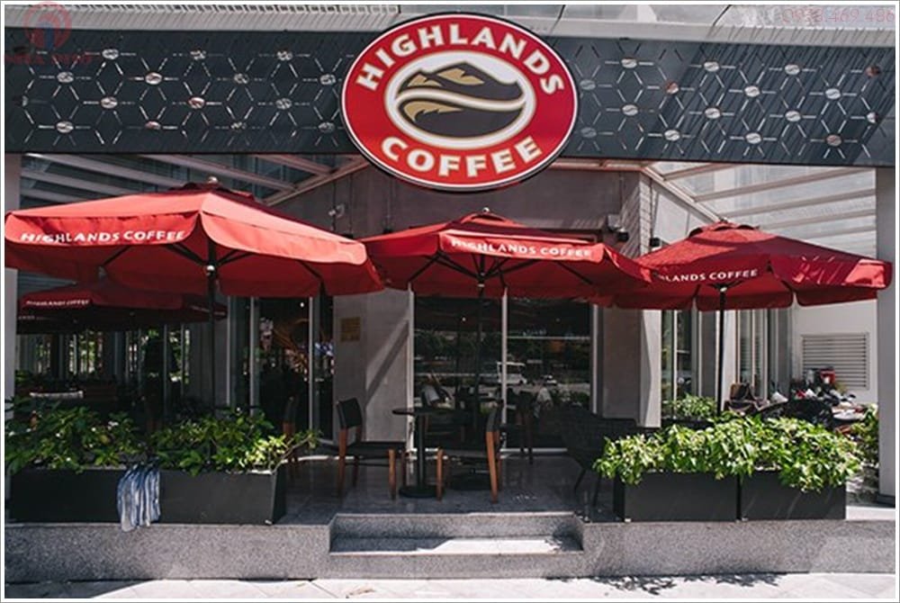Top 32 thương hiệu nhượng quyền cafe siêu lợi nhuận 21 Quán cà phê với logo lớn phía trên, dù đỏ bên ngoài và cây xanh trang trí phía trước.