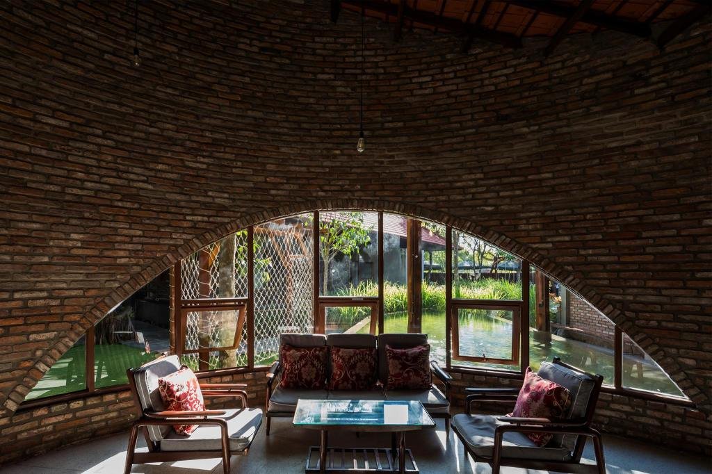 Kh&ocirc;ng gian nội thất với tường gạch cong, b&agrave;n ghế gỗ, cửa sổ nh&igrave;n ra s&acirc;n vườn xanh m&aacute;t v&agrave; &aacute;nh s&aacute;ng tự nhi&ecirc;n.