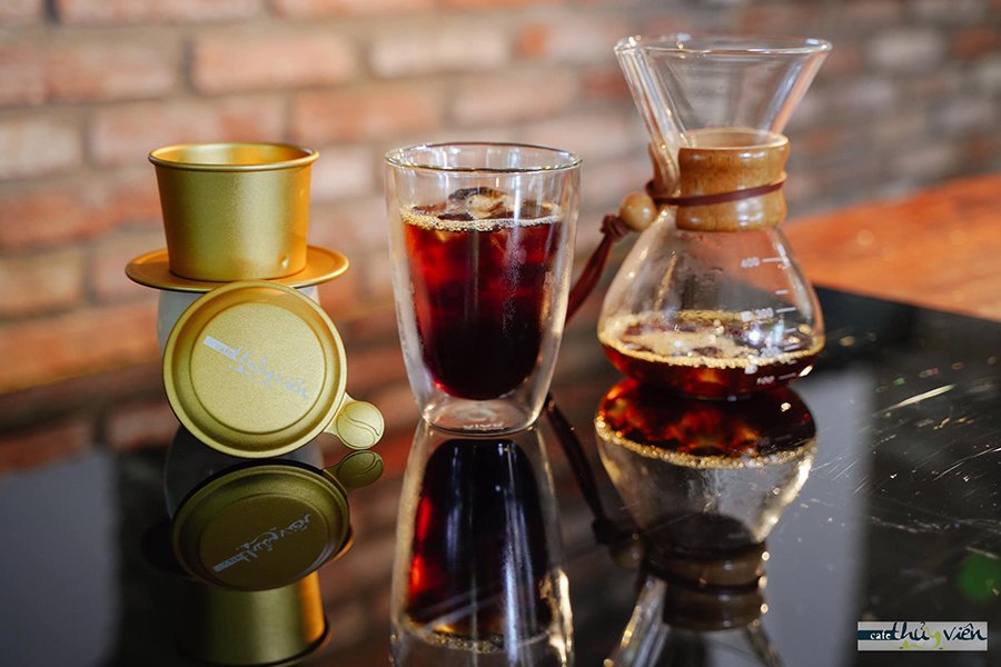 Ly c&agrave; ph&ecirc; đ&aacute;, b&igrave;nh pha v&agrave; phin c&agrave; ph&ecirc; tr&ecirc;n b&agrave;n k&iacute;nh, nền gạch đỏ mờ.