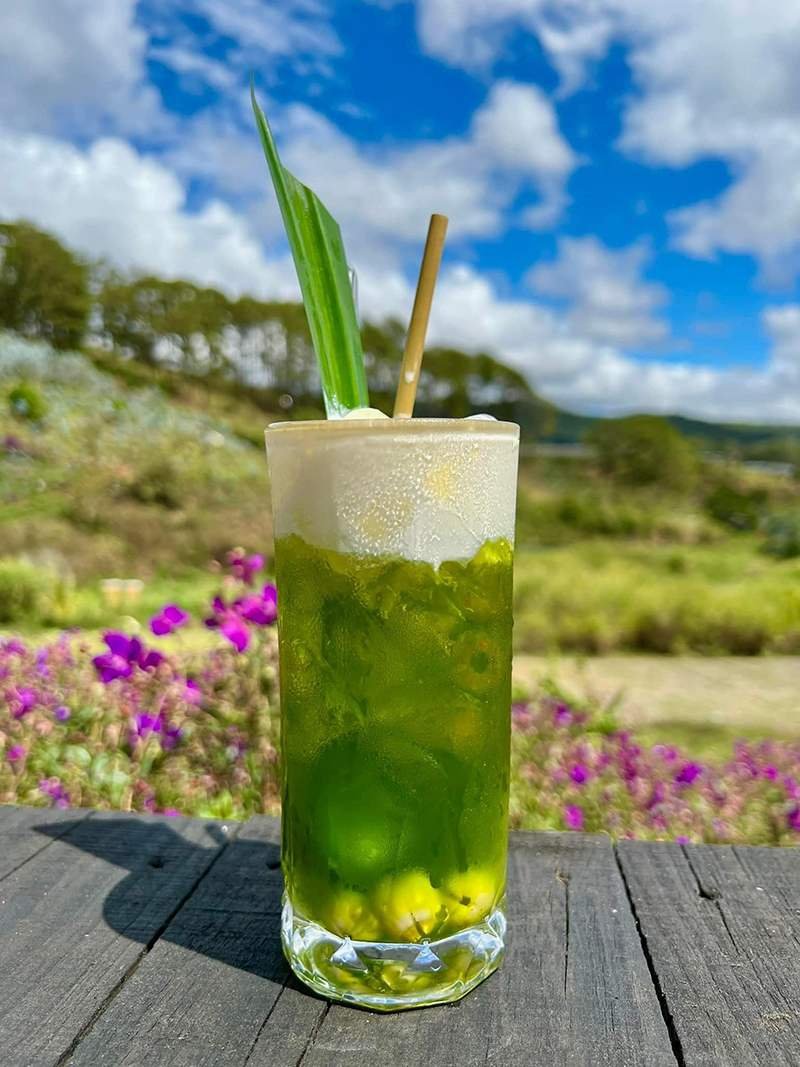 Ly nước m&agrave;u xanh với l&aacute; trang tr&iacute;, đặt tr&ecirc;n b&agrave;n gỗ ngo&agrave;i trời, hậu cảnh l&agrave; bầu trời xanh v&agrave; c&acirc;y cỏ.