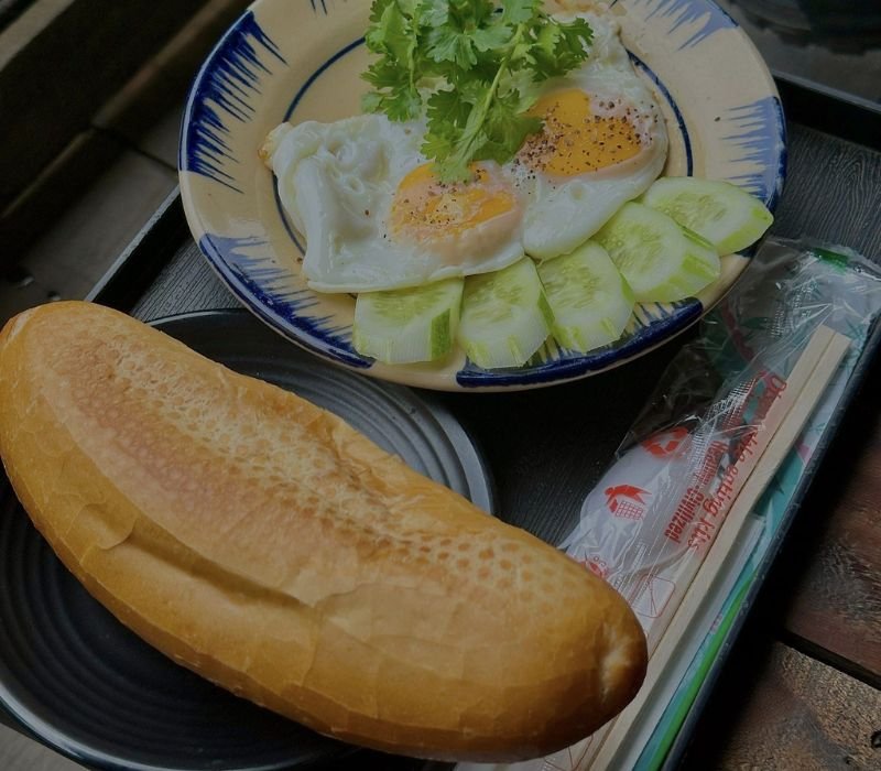 Đĩa trứng ốp la với rau m&ugrave;i v&agrave; dưa leo, b&ecirc;n cạnh l&agrave; một ổ b&aacute;nh m&igrave;, đũa v&agrave; gia vị được b&agrave;y tr&ecirc;n b&agrave;n.