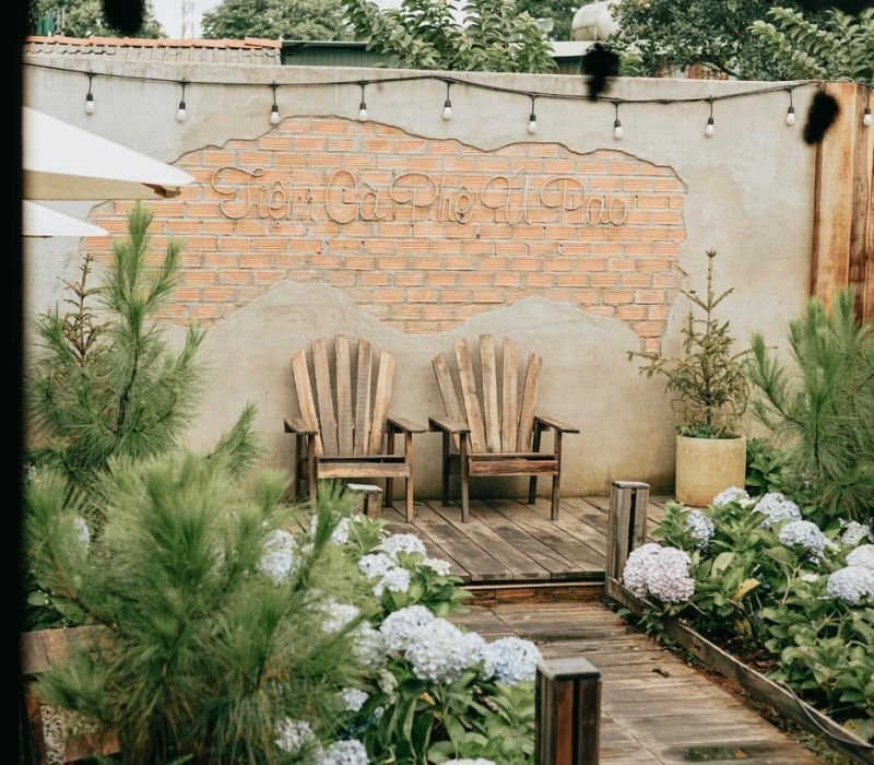 Hai ghế gỗ đặt tr&ecirc;n s&agrave;n gỗ trước bức tường gạch, xung quanh l&agrave; c&acirc;y xanh v&agrave; hoa cẩm t&uacute; cầu.