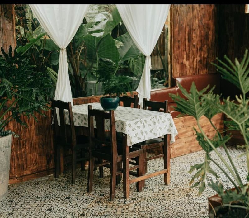 B&agrave;n ghế gỗ với khăn trải b&agrave;n trắng, xung quanh l&agrave; c&acirc;y xanh v&agrave; r&egrave;m trắng trong kh&ocirc;ng gian nội thất ấm c&uacute;ng.