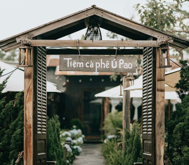 Cổng gỗ với biển hiệu "Tiệm c&agrave; ph&ecirc; &Uacute; Pao", xung quanh c&oacute; c&acirc;y xanh v&agrave; &ocirc; che nắng.