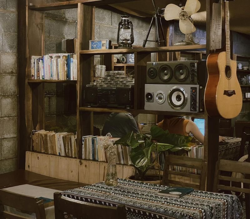Qu&aacute;n c&agrave; ph&ecirc; ấm c&uacute;ng với kệ s&aacute;ch, loa, đ&agrave;n guitar treo tường v&agrave; b&agrave;n ghế gỗ, tạo kh&ocirc;ng gian thư gi&atilde;n.