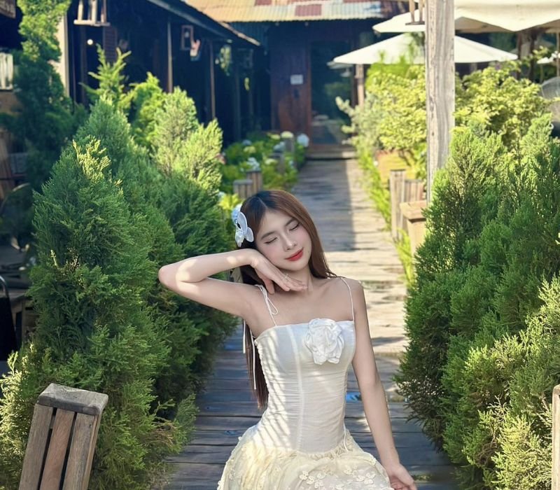 Người phụ nữ mặc v&aacute;y trắng đứng tr&ecirc;n lối đi bằng gỗ, xung quanh l&agrave; c&acirc;y xanh v&agrave; c&aacute;c t&ograve;a nh&agrave; nhỏ.