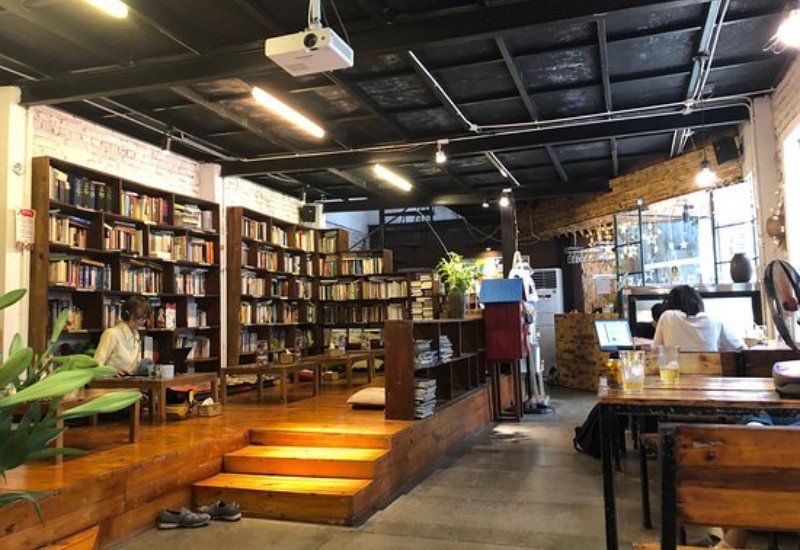 Qu&aacute;n c&agrave; ph&ecirc; với kệ s&aacute;ch lớn, b&agrave;n ghế gỗ, hai người đang ngồi l&agrave;m việc, kh&ocirc;ng gian ấm c&uacute;ng v&agrave; thư gi&atilde;n.