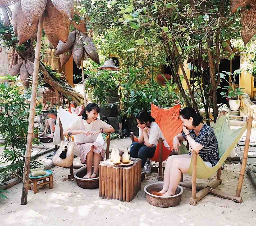 Ba người thư gi&atilde;n ng&acirc;m ch&acirc;n trong bồn gỗ tại một khu vườn c&oacute; c&acirc;y cối xanh m&aacute;t v&agrave; ghế tre.