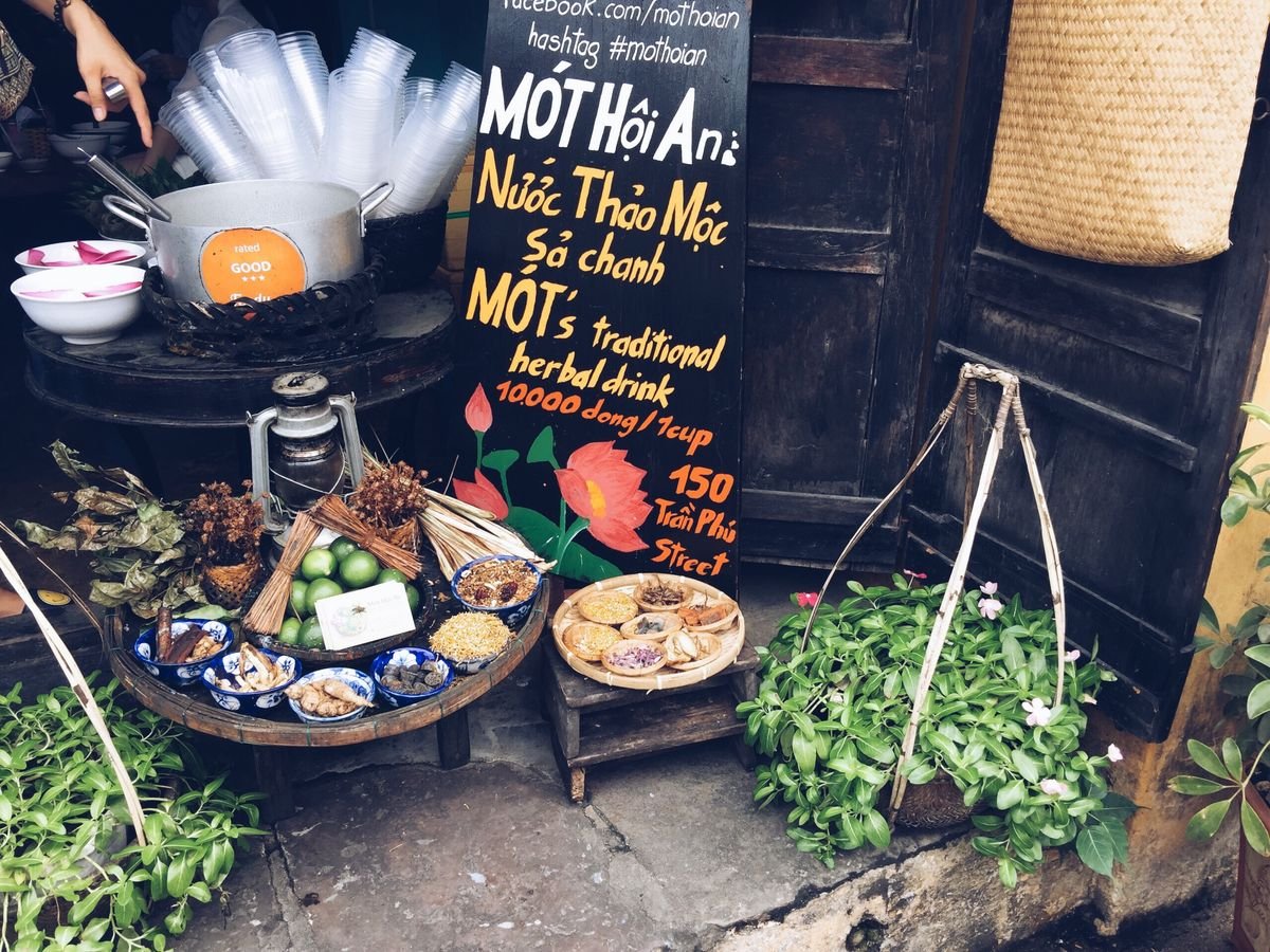 Quầy b&aacute;n nước thảo mộc với bảng hiệu, ly nhựa v&agrave; c&aacute;c nguy&ecirc;n liệu thảo mộc b&agrave;y tr&iacute; tr&ecirc;n b&agrave;n gỗ.