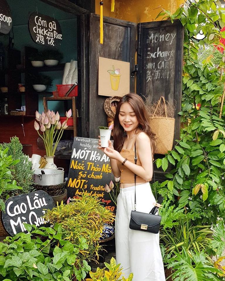 Ảnh: @joly.mi Người đứng trước quán nhỏ với bảng hiệu và cây xanh xung quanh, cầm một ly nước thảo mộc.