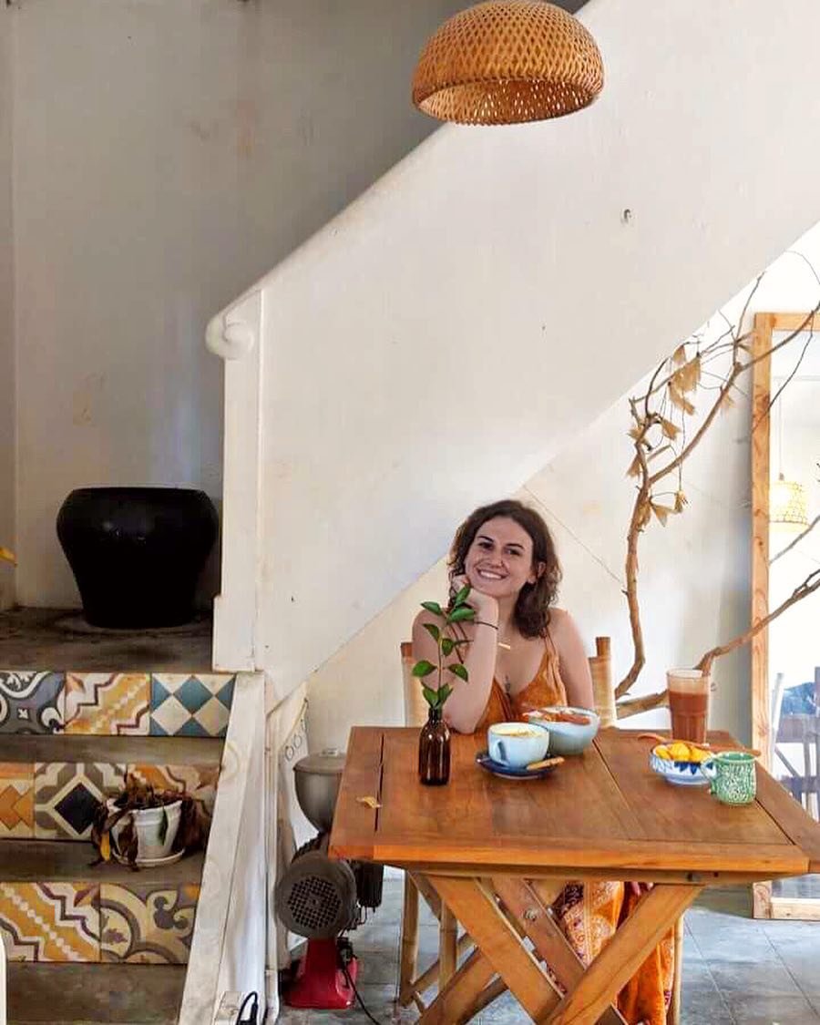 Ảnh: @alysescotti Người phụ nữ ngồi tại bàn gỗ nhỏ, bên cầu thang trang trí gạch hoa, với cốc và đĩa trên bàn cạnh chậu cây.