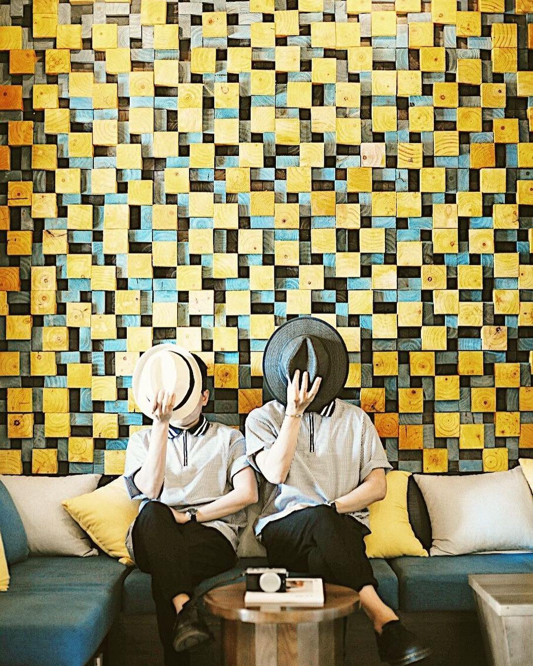 Ảnh: @fusioncafehoian Hai người ngồi trên ghế, che mặt bằng nón, trước bức tường họa tiết ô vuông màu vàng và xanh.