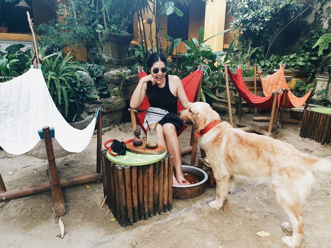 Ảnh: @qq_1997_qq Người ngồi trên ghế ngoài trời cạnh bàn tròn, chân ngâm trong chậu nước, bên cạnh là một chú chó lông vàng.