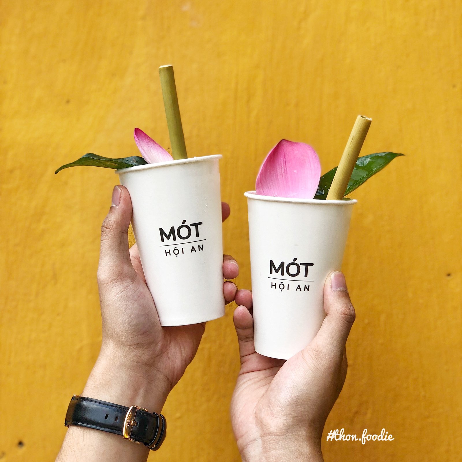 Ảnh: @thon.foodie Hai ly nước Mót Hội An đang được cầm trên tay, trang trí bằng lá và cánh hoa sen, nền màu vàng.
