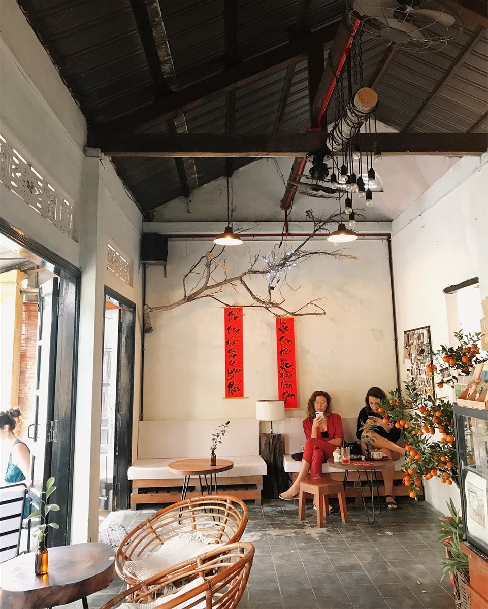 Ảnh: @dulichhoian Quán cà phê nhỏ với bàn ghế gỗ, trang trí cành cây và câu đối đỏ trên tường, hai người đang ngồi thư giãn.