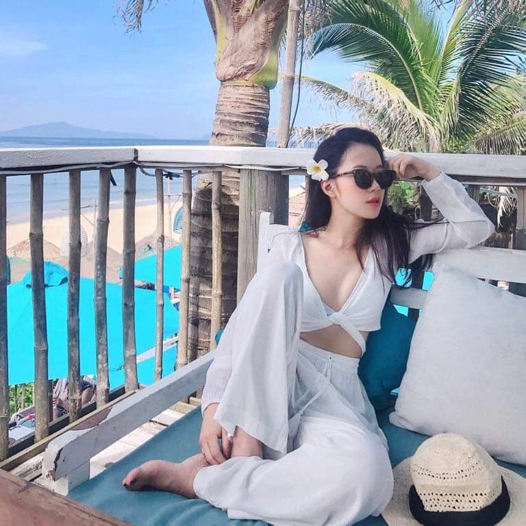 Ảnh: @iam.vtt12 Người phụ nữ mặc đồ trắng ngồi thư giãn trên ghế ngoài trời, phía sau là bãi biển và cây dừa.