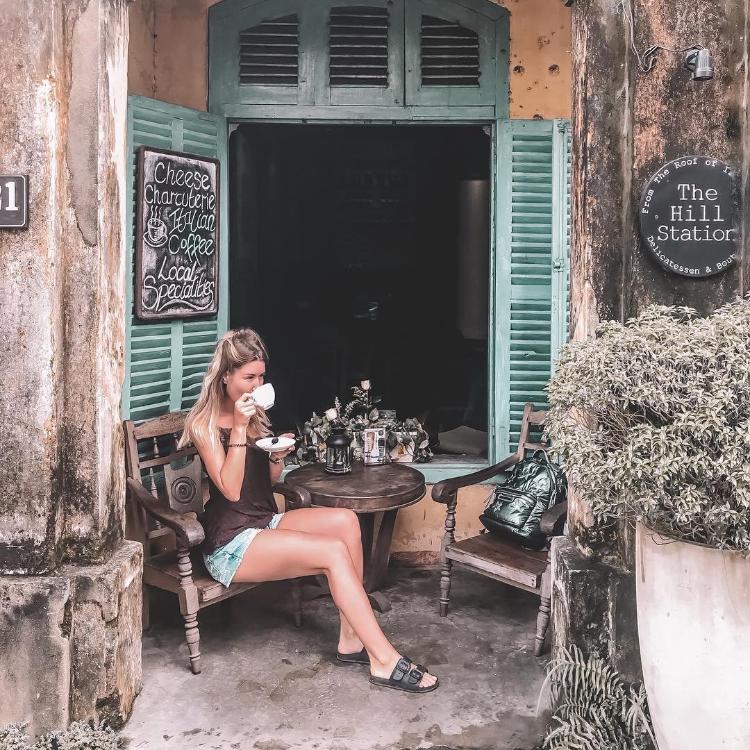 Ảnh: @sashaa_pashaa Người phụ nữ ngồi uống cà phê trước quán với cửa sổ xanh, bảng thực đơn và chậu cây lớn.