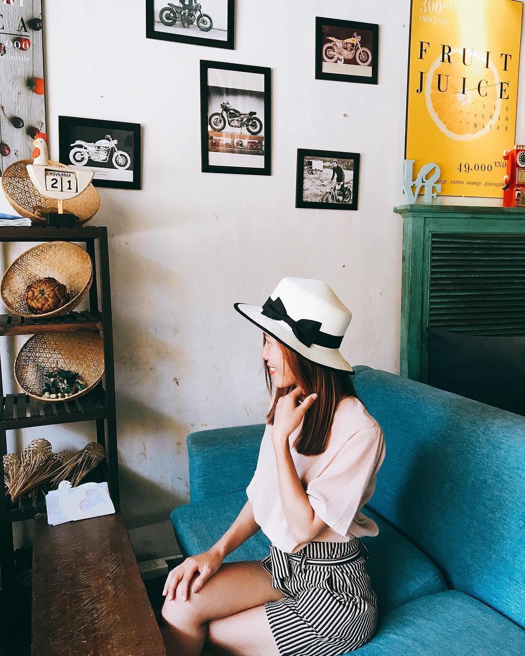 Ảnh: @uyenmin.96 Người phụ nữ đội mũ ngồi trên ghế xanh, bên cạnh là kệ trang trí và bức tường treo ảnh xe máy.
