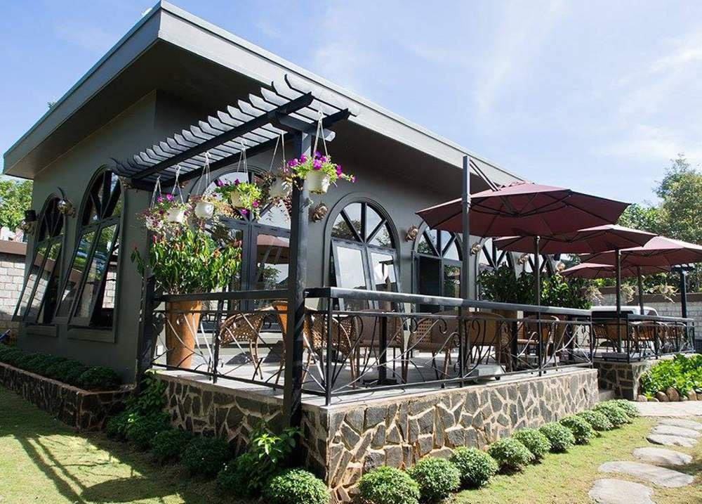 Quán cà phê với thiết kế hiện đại, có mái che và bàn ghế ngoài trời, xung quanh là cây xanh và hoa treo.