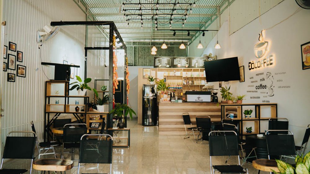 Quán cà phê với không gian hiện đại, bàn ghế gỗ và kim loại, trang trí cây xanh, quầy pha chế ở phía sau.