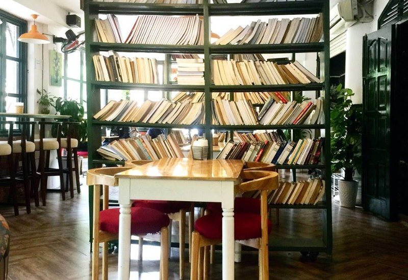 Kệ s&aacute;ch với nhiều s&aacute;ch được sắp xếp, ph&iacute;a trước l&agrave; b&agrave;n v&agrave; ghế trong một kh&ocirc;ng gian qu&aacute;n c&agrave; ph&ecirc; tho&aacute;ng đ&atilde;ng.