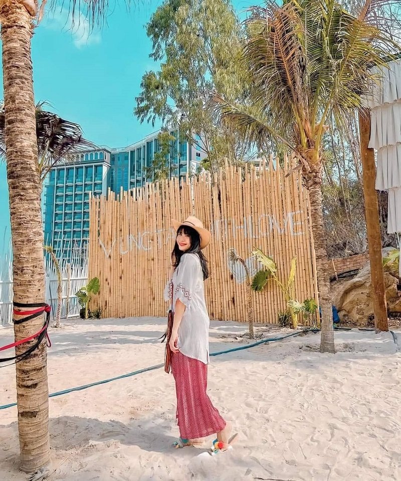 Người phụ nữ đội mũ đứng trên bãi cát, phía sau là hàng rào tre và cây cọ, một tòa nhà cao tầng phía xa.