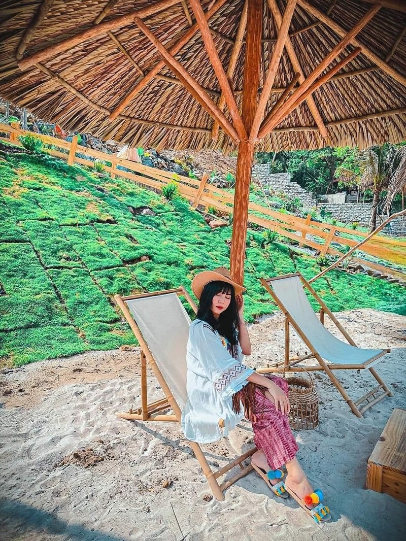 Người ngồi trên ghế dưới dù lá tại bãi biển, xung quanh có cỏ xanh và hàng rào gỗ.