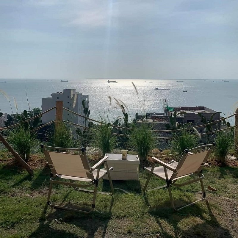 Hai ghế và bàn nhỏ trên bãi cỏ, nhìn ra biển với tàu thuyền xa xa dưới ánh nắng mặt trời.