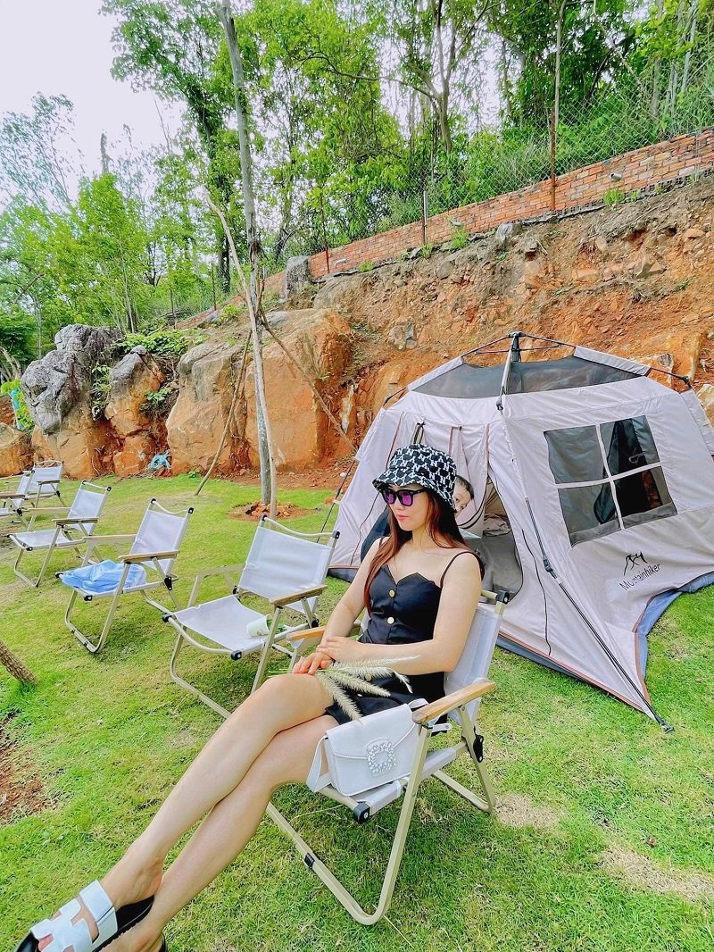 Người ngồi trên ghế gấp ngoài trời, cạnh lều trại, trên bãi cỏ xanh, xung quanh là cây cối và vách đá.