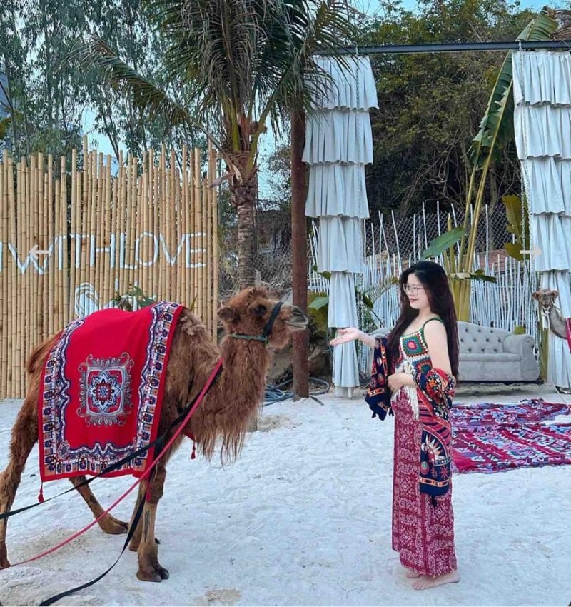 Người phụ nữ trong trang phục họa tiết đứng cạnh lạc đà trên nền cát trắng, phía sau là cây cối và rèm trang trí.