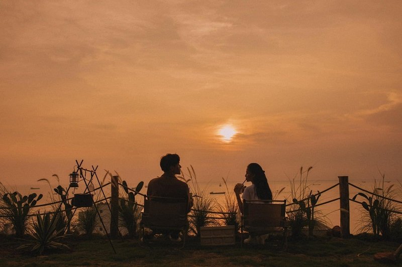 Hai người ngồi trên ghế ngoài trời, ngắm hoàng hôn trên biển, xung quanh là thiên nhiên và cây cỏ.
