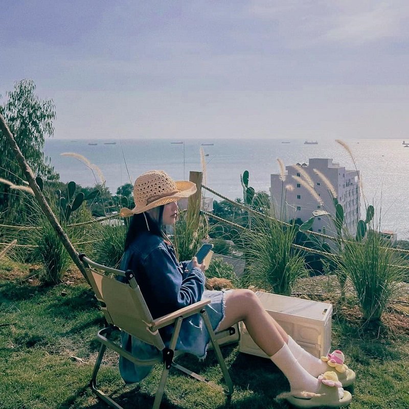 Người ngồi trên ghế gấp ngoài trời, đội mũ rơm, cầm điện thoại, phông nền biển và tàu xa xa.