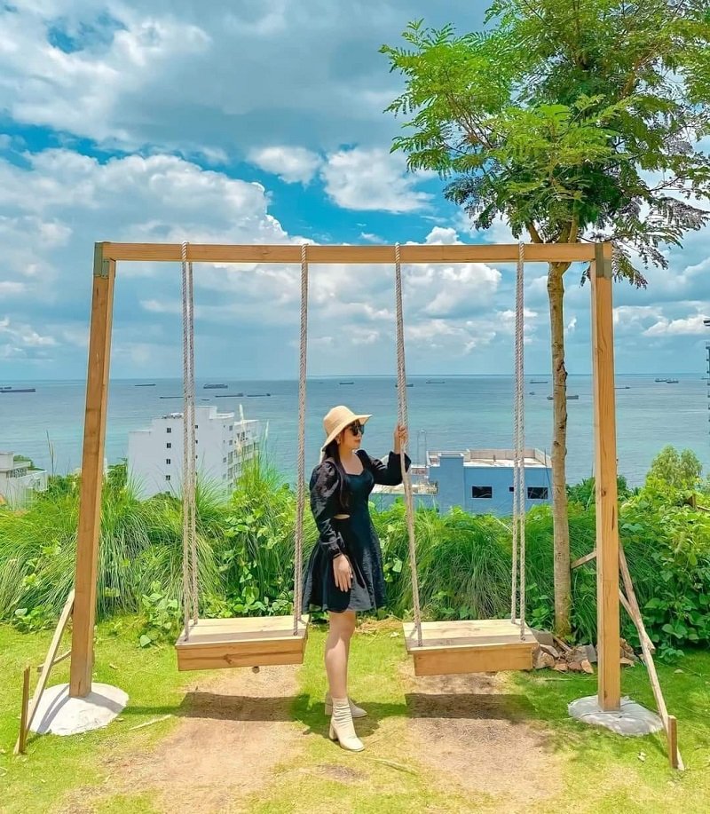 Người đội mũ đứng cạnh xích đu gỗ, phía sau là biển xanh và bầu trời nhiều mây.