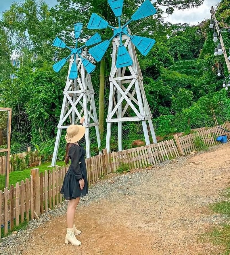 Người phụ nữ đội mũ đứng trước cối xay gió màu xanh, xung quanh là hàng rào và cây xanh.