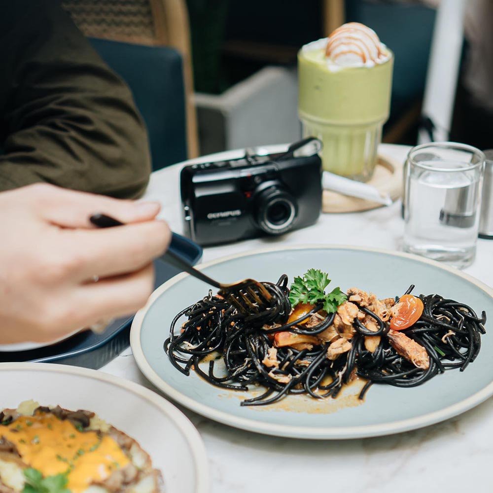 Đĩa m&igrave; &yacute; mực, tay cầm nĩa, b&ecirc;n cạnh l&agrave; m&aacute;y ảnh, ly nước xanh v&agrave; ly nước lọc tr&ecirc;n b&agrave;n.