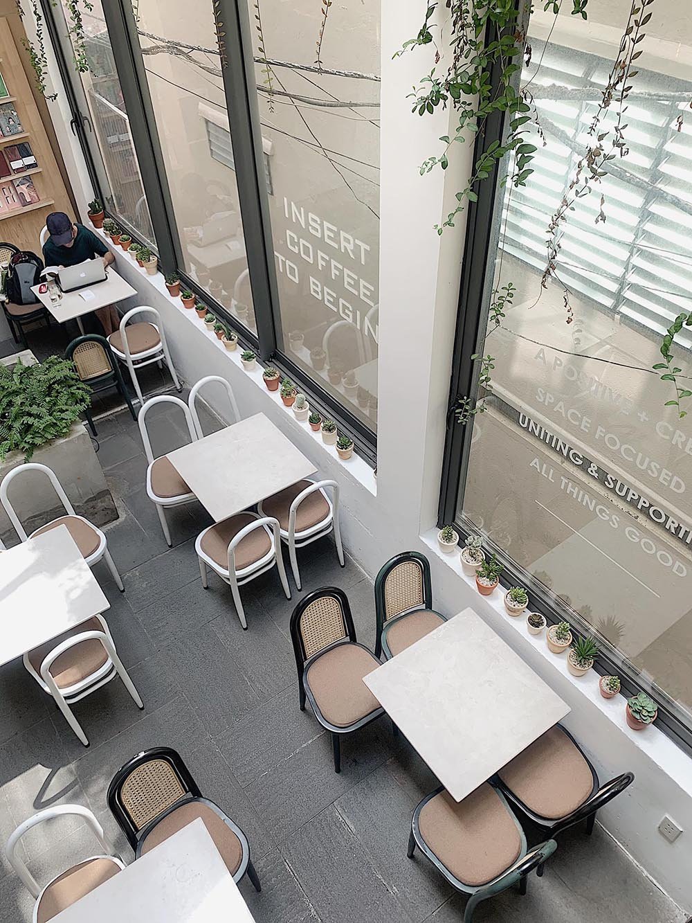 Qu&aacute;n c&agrave; ph&ecirc; với b&agrave;n ghế sắp xếp ngăn nắp, cửa sổ lớn v&agrave; c&acirc;y xanh trang tr&iacute;, một người l&agrave;m việc với laptop.