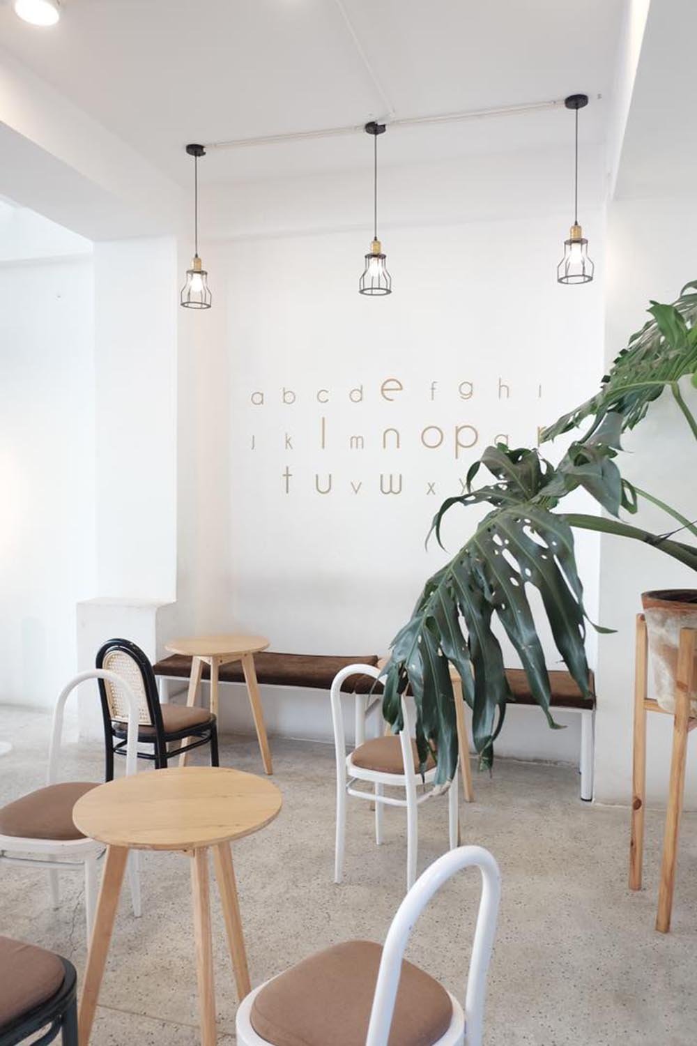 Qu&aacute;n c&agrave; ph&ecirc; với b&agrave;n ghế gỗ, c&acirc;y xanh, tường trắng trang tr&iacute; chữ c&aacute;i, đ&egrave;n treo trần phong c&aacute;ch hiện đại.