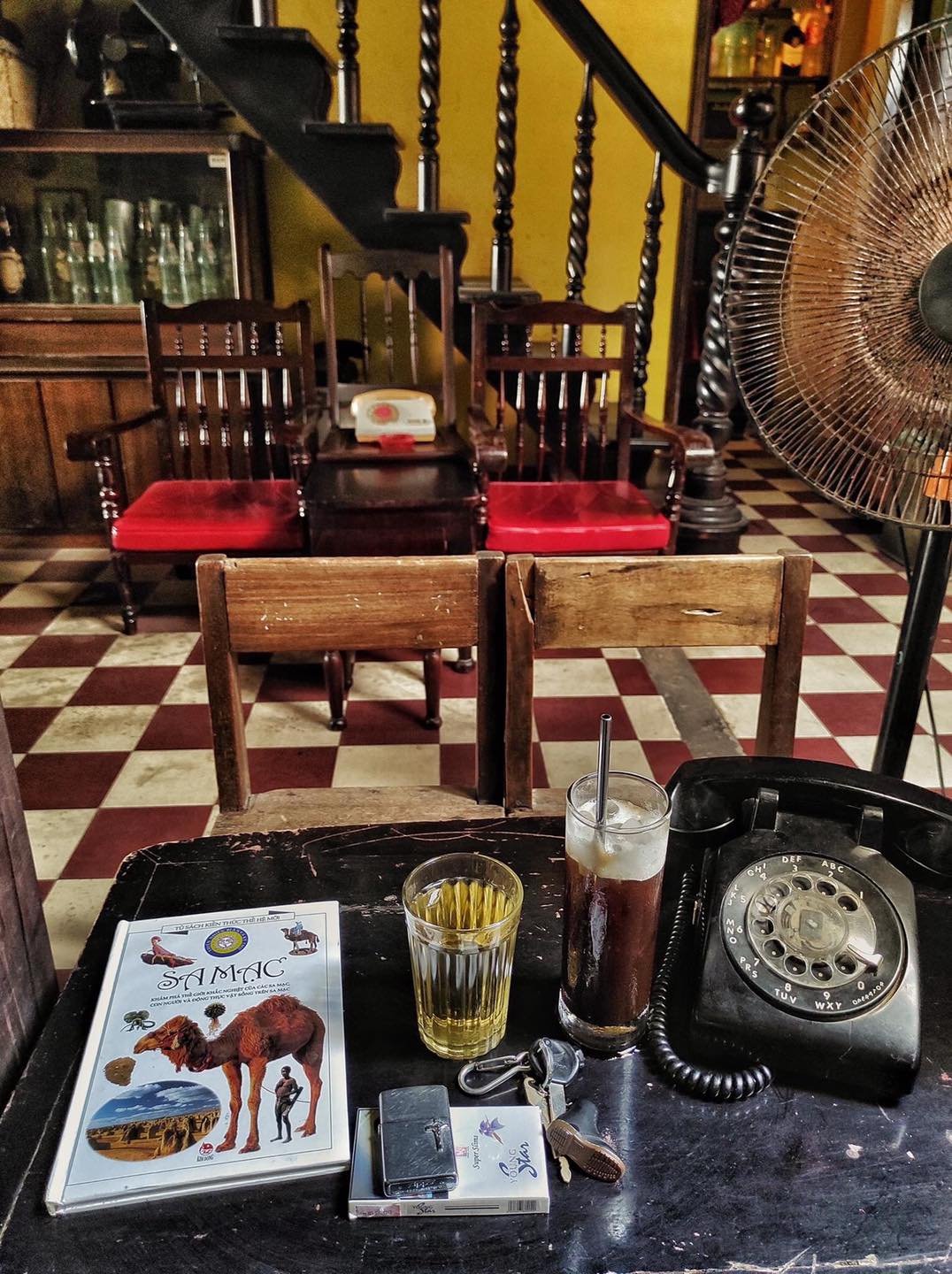 B&agrave;n gỗ với s&aacute;ch, ly nước, ch&igrave;a kh&oacute;a, m&aacute;y quay số cổ điển trong kh&ocirc;ng gian nội thất cổ điển với ghế gỗ v&agrave; quạt điện.