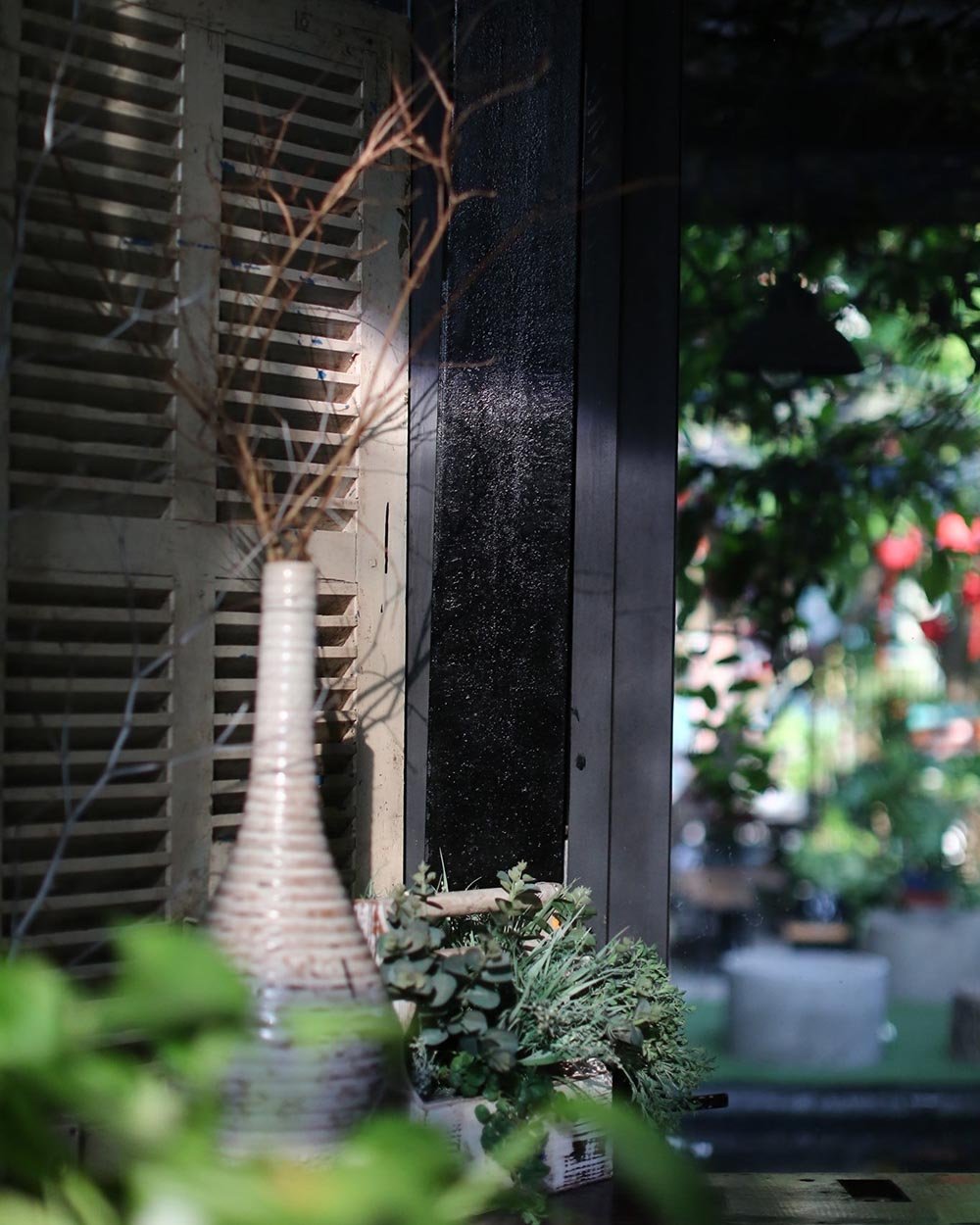 B&igrave;nh gốm trắng với c&agrave;nh kh&ocirc;, đặt tr&ecirc;n b&agrave;n cạnh cửa sổ, &aacute;nh s&aacute;ng chiếu qua, nền c&acirc;y xanh mờ ph&iacute;a sau.