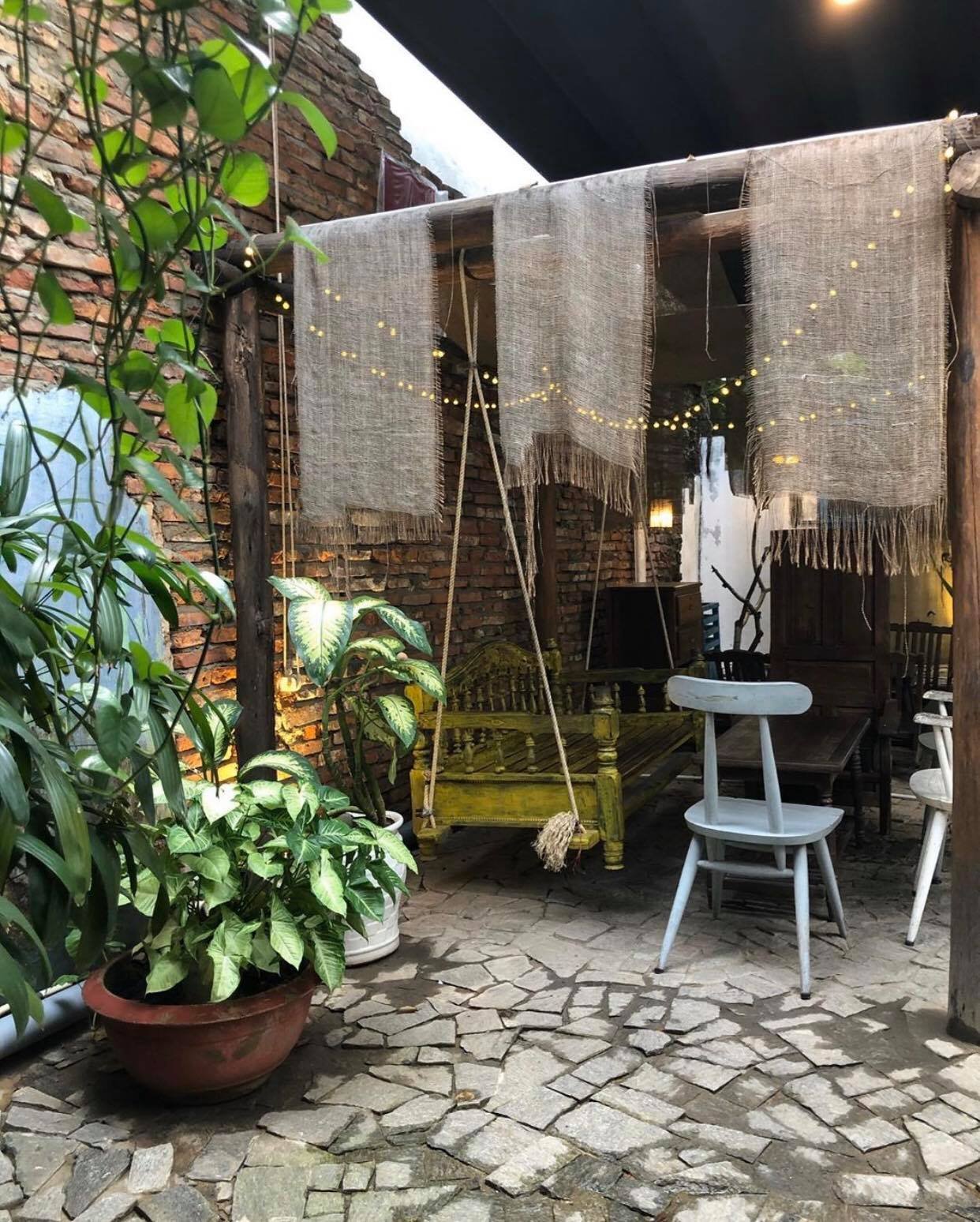 Khu vườn nhỏ với ghế gỗ, c&acirc;y xanh v&agrave; đ&egrave;n trang tr&iacute; treo dưới tấm vải, tạo kh&ocirc;ng gian thư gi&atilde;n ấm c&uacute;ng.