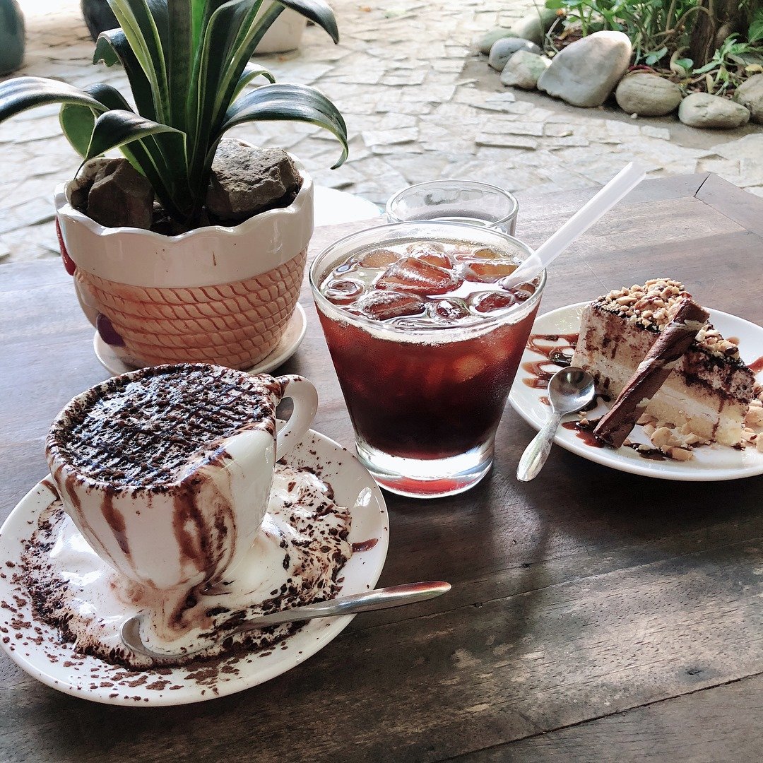 Ly c&agrave; ph&ecirc; đ&aacute;, t&aacute;ch c&agrave; ph&ecirc; sữa n&oacute;ng v&agrave; đĩa b&aacute;nh ngọt tr&ecirc;n b&agrave;n gỗ cạnh chậu c&acirc;y trang tr&iacute;.