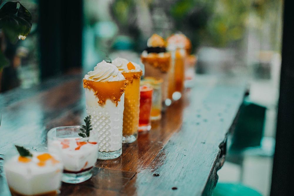 Nhiều ly đồ uống v&agrave; m&oacute;n tr&aacute;ng miệng trang tr&iacute; đẹp mắt xếp h&agrave;ng tr&ecirc;n b&agrave;n gỗ trong kh&ocirc;ng gian tho&aacute;ng đ&atilde;ng.