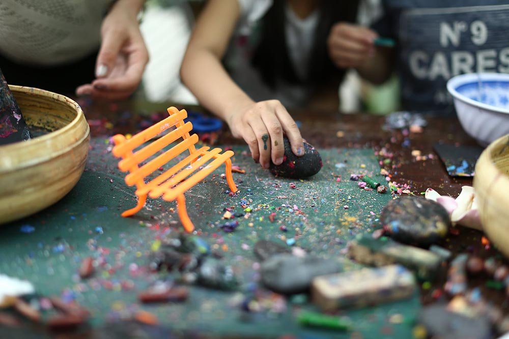 B&agrave;n tay trẻ em t&ocirc; m&agrave;u l&ecirc;n sỏi, xung quanh l&agrave; dụng cụ vẽ v&agrave; m&ocirc; h&igrave;nh ghế nhỏ m&agrave;u cam, tạo kh&ocirc;ng gian s&aacute;ng tạo.