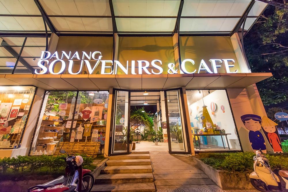 Mặt tiền qu&aacute;n c&agrave; ph&ecirc; v&agrave; cửa h&agrave;ng qu&agrave; lưu niệm với bảng hiệu s&aacute;ng đ&egrave;n v&agrave;o ban đ&ecirc;m, c&oacute; xe m&aacute;y đỗ ph&iacute;a trước.